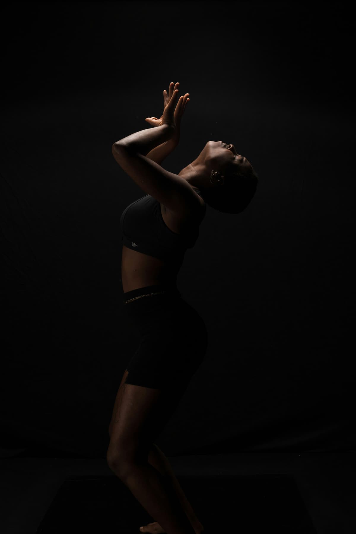 Black woman in black training wear arching backward, hands raised, rim-lit against black.